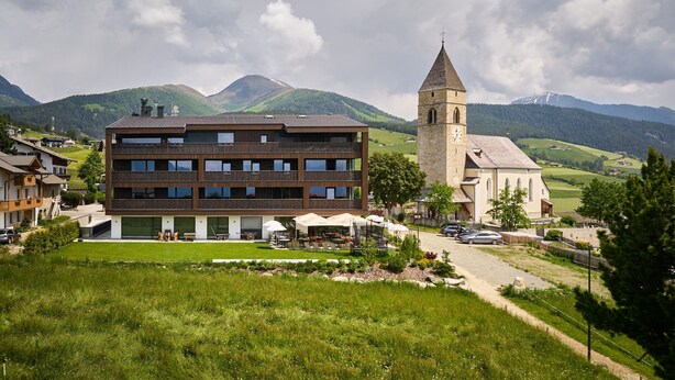 A modern building with a large windowed front, a church with a bell tower, and a mountainous backdrop.