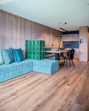 A modern living room with a sectional sofa, a green modular seating unit, and a kitchen area with wooden walls and a built-in oven.