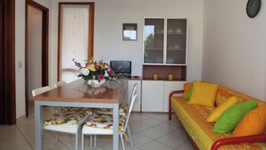 A small, well-lit room with a dining table, chairs, a sofa, a TV, and a window with curtains. - Apartment in a Villa With Garden (San Michele al Tagliamento)