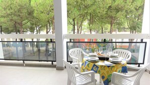 Un balcon avec une table dressée pour le dîner, entouré de chaises blanches, donnant sur une rue bordée d’arbres.
