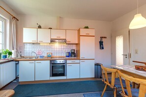 Kompakti keittiö, jossa on valkoiset kaapit, sisäänrakennettu uuni, pesuallas sekä ruokailutila, jossa on pöytä ja tuolit.