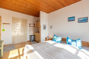 A bedroom with a bed, a wooden ceiling, a door, a small kitchen area, and two framed pictures on the wall.