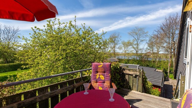 Una terraza con un paraguas rojo, una silla con cojín floral y vista a un lago.