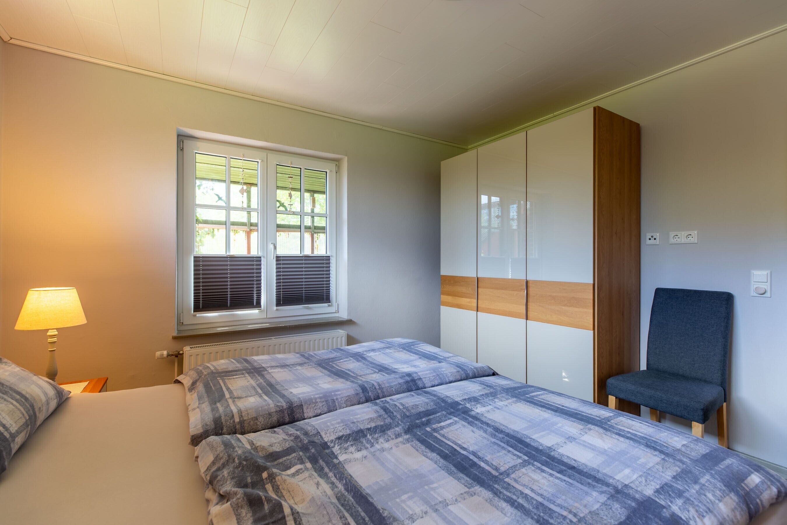 A bedroom with two beds, a window with blinds, a chair, and a wardrobe.