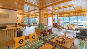 Una habitación espaciosa con suelo de madera, un área de estar central, una mesa de madera con piezas de ajedrez y grandes ventanales con vistas al exterior.