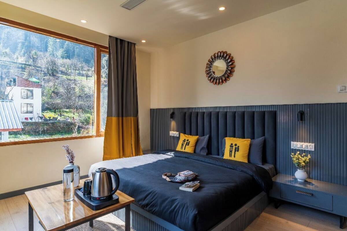 Une chambre moderne avec un grand lit, une table de chevet, une commode, une fenêtre avec des rideaux et une vue sur un immeuble et des arbres.