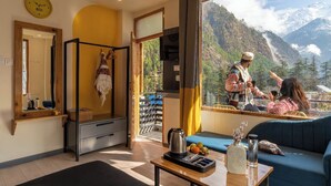 Una habitación con un ventanal grande que da a un paisaje montañoso, una cama, una mesa con hervidor y frutas, y un espejo en la pared.
