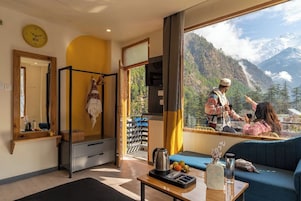 A room with a large window overlooking a mountainous landscape, a bed, a table with a kettle and fruits, and a mirror on the wall.