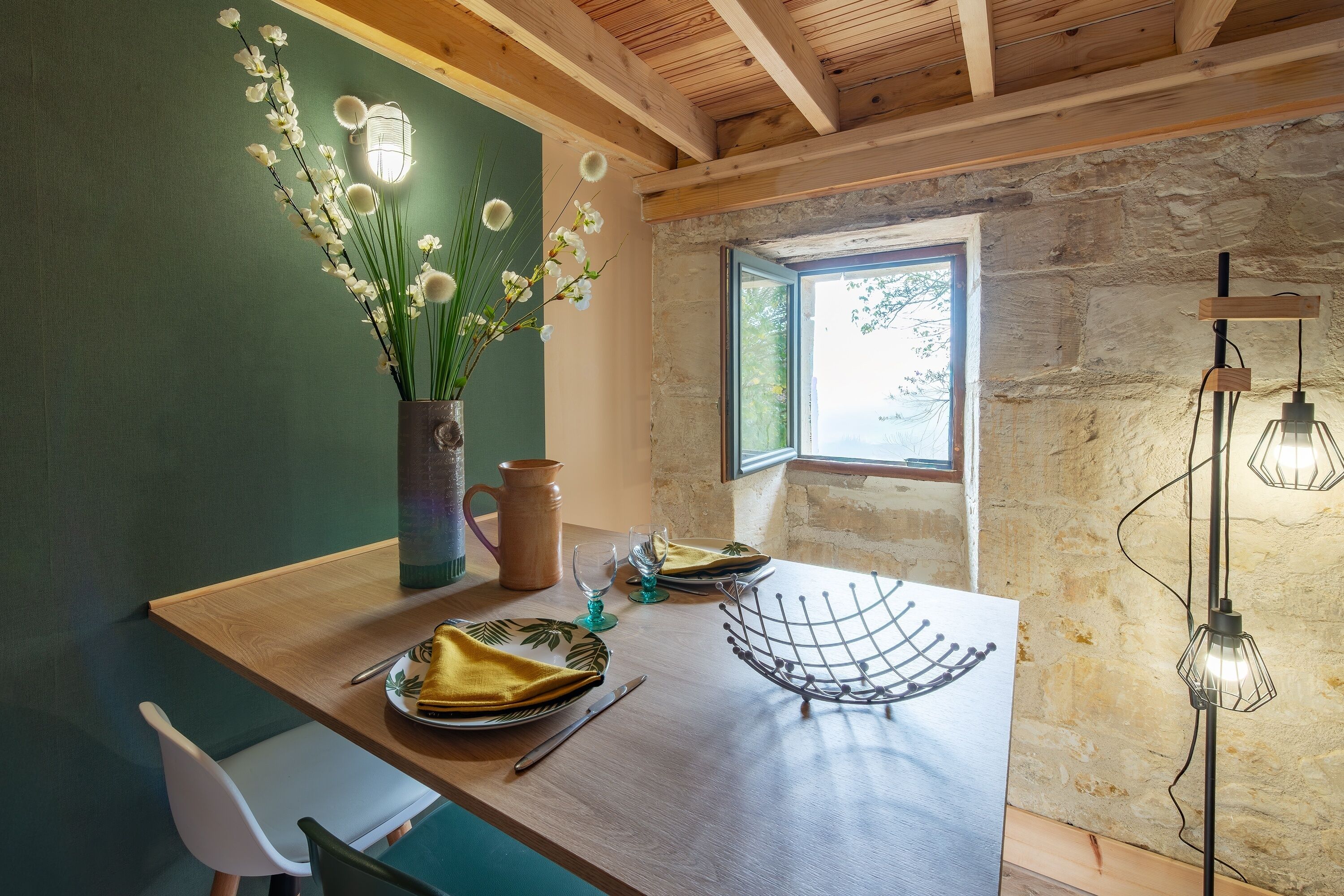 Une table de salle à manger en bois, ornée d’un vase de fleurs blanches, d’une tasse en cuivre et d’un panier en fil métallique.