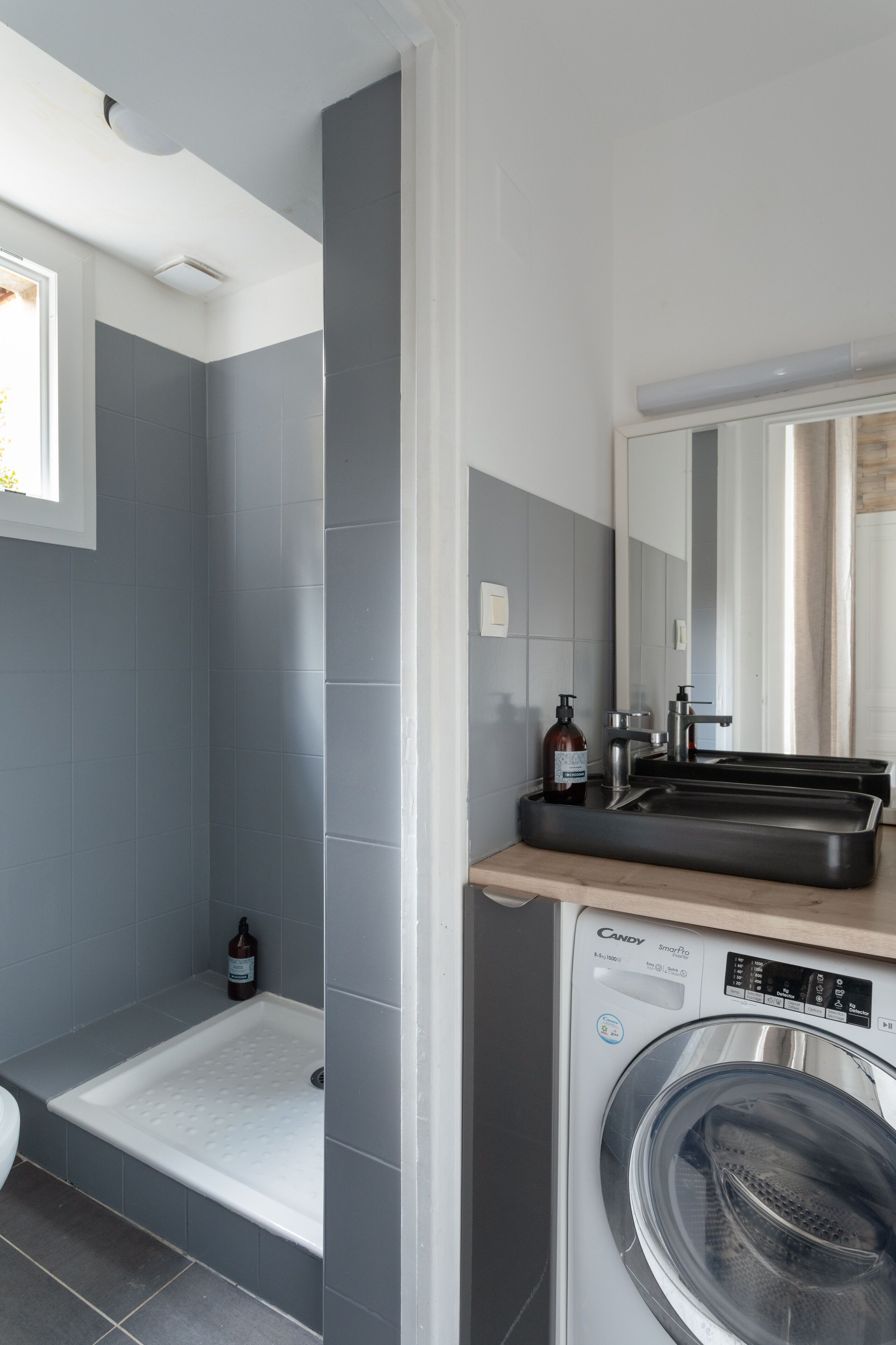 Une salle de bain moderne avec des carreaux gris, un lave-linge encastré, un lavabo avec un miroir et un espace douche.