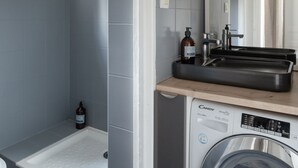 A modern bathroom with grey tiles, a built-in washing machine, a sink with a mirror, and a shower area. - Le Petit 18 - T2 proche de la gare (Rennes)