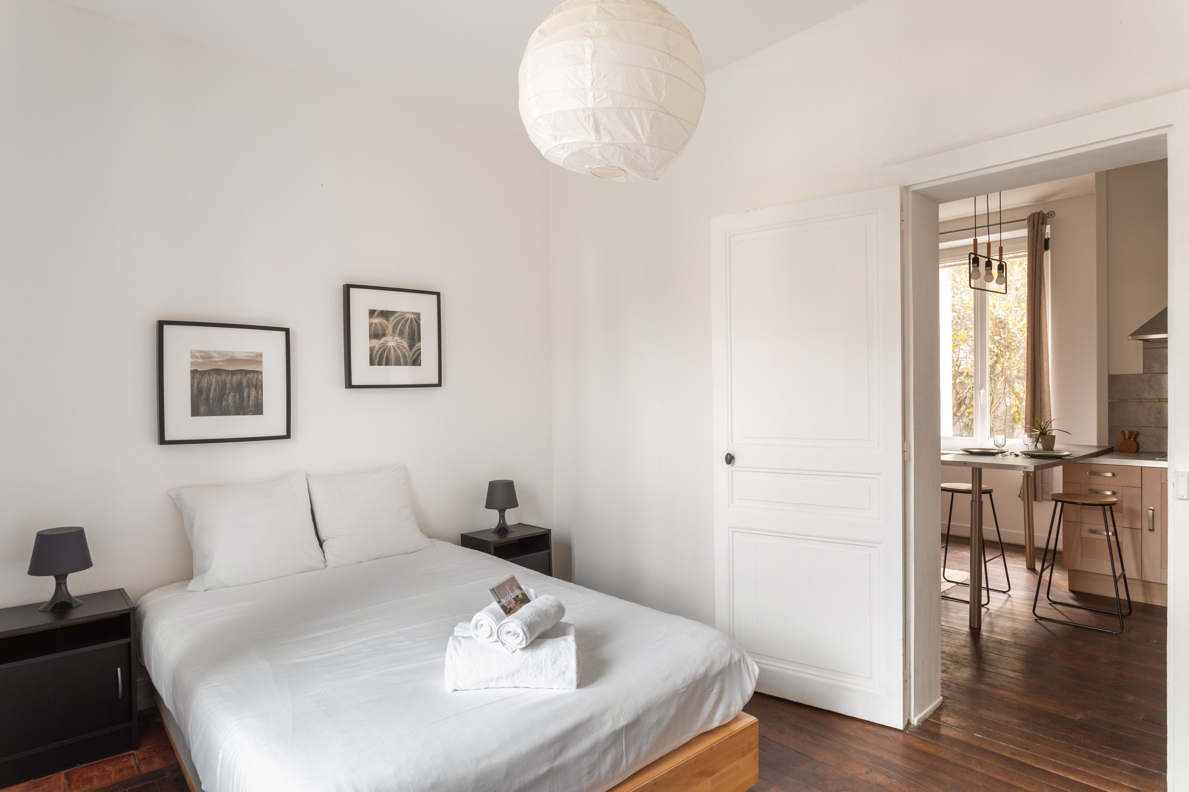 Une chambre à coucher comprenant un lit, des tables de chevet, des tableaux encadrés et une porte donnant sur un coin cuisine.