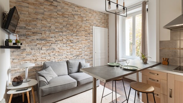 A modern kitchen with a stone wall, a grey sofa, and a dining area with stools. - Le Petit 18 - T2 proche de la gare (Rennes)