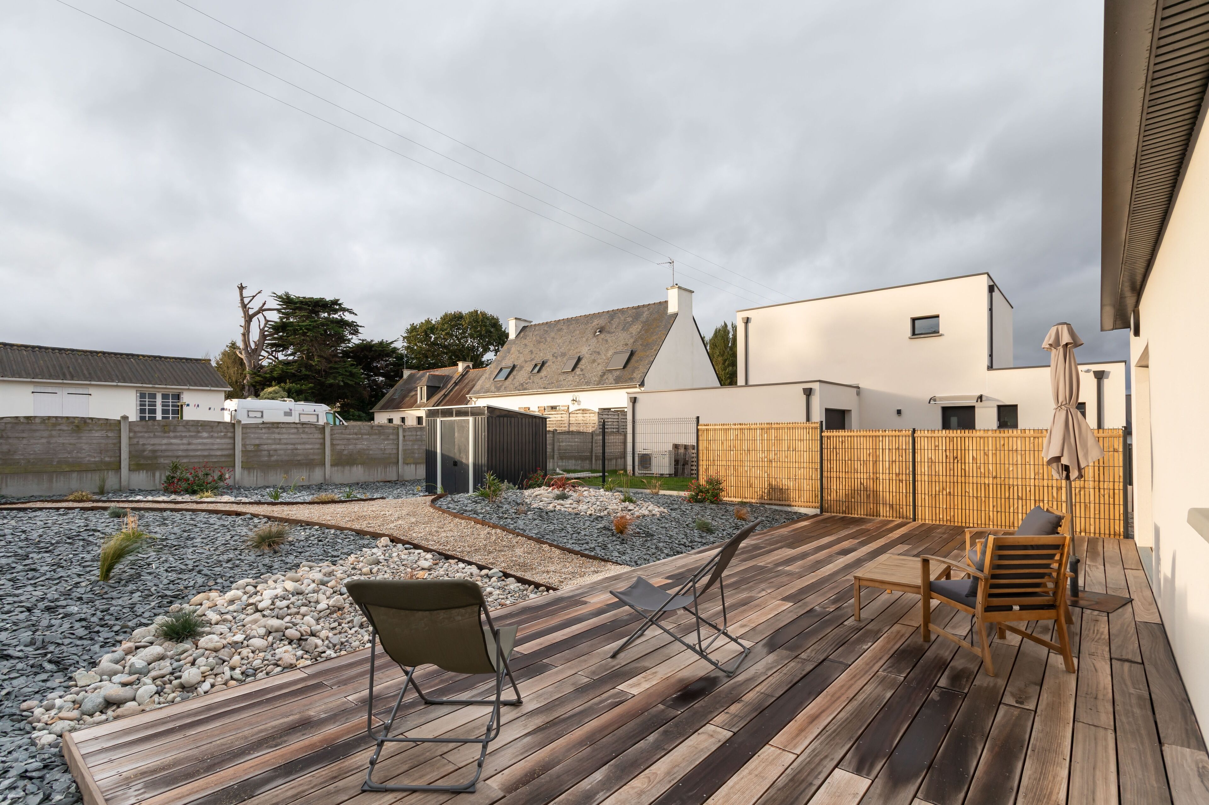 Une terrasse en bois avec deux chaises, un petit jardin agrémenté de rochers et de plantes, et un bâtiment blanc avec un toit gris en arrière-plan.