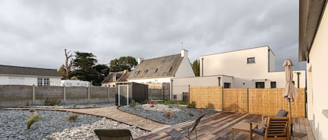 Une terrasse en bois avec deux chaises, un petit jardin agrémenté de rochers et de plantes, et un bâtiment blanc avec un toit gris en arrière-plan.