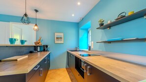 Eine moderne Küche mit Holzarbeitsplatten, einem eingebauten Backofen und einem Spülbecken. Zwei Pendelleuchten hängen von der Decke, an der Wand hängt ein gerahmtes Bild und ein Regal ist mit Küchenutensilien bestückt.