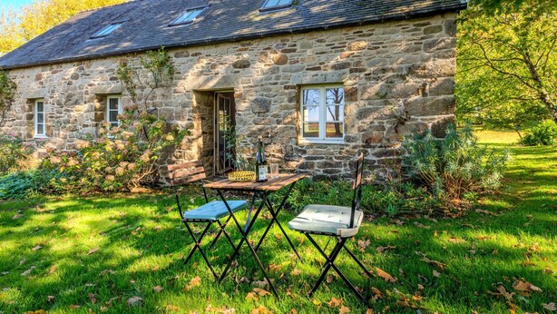 Ein Steinhäuschen mit schrägem Dach, zwei Fenstern und einer Tür. Vor dem Haus befindet sich ein kleiner Garten mit Rasen und ein Tisch mit Stühlen.