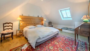 Ein Schlafzimmer mit einem hölzernen Kopfteil, einem Bett mit weißer Bettwäsche, einem Teppich mit rotem Muster, einem Nachttisch aus Holz, einem Sessel, einem kleinen Tisch mit einer Lampe und einem Fenster mit Blick.