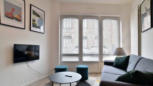Una sala de estar moderna con un sofá gris, una mesa de centro redonda y dos otomanas azules. Hay un televisor de pantalla plana montado en la pared, y dos ventanas con persianas.