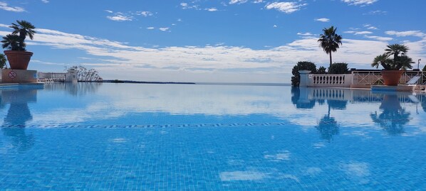 Una piscina azul cristalina con borde de azulejos, palmeras y una vista distante del mar bajo un cielo parcialmente nublado.