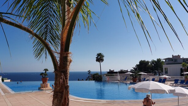 Zona de piscina con sillones de descanso y sombrillas, una palmera y vista al mar.