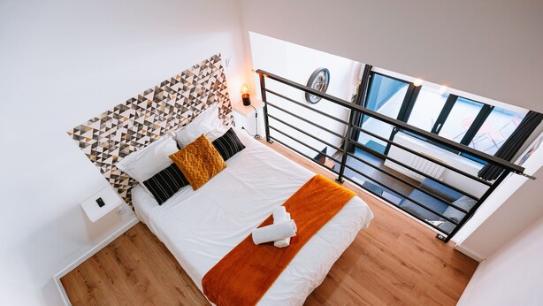 A modern bedroom with a wooden floor, a bed with white bedding and a brown throw, a geometric-patterned headboard, and a balcony with a railing.