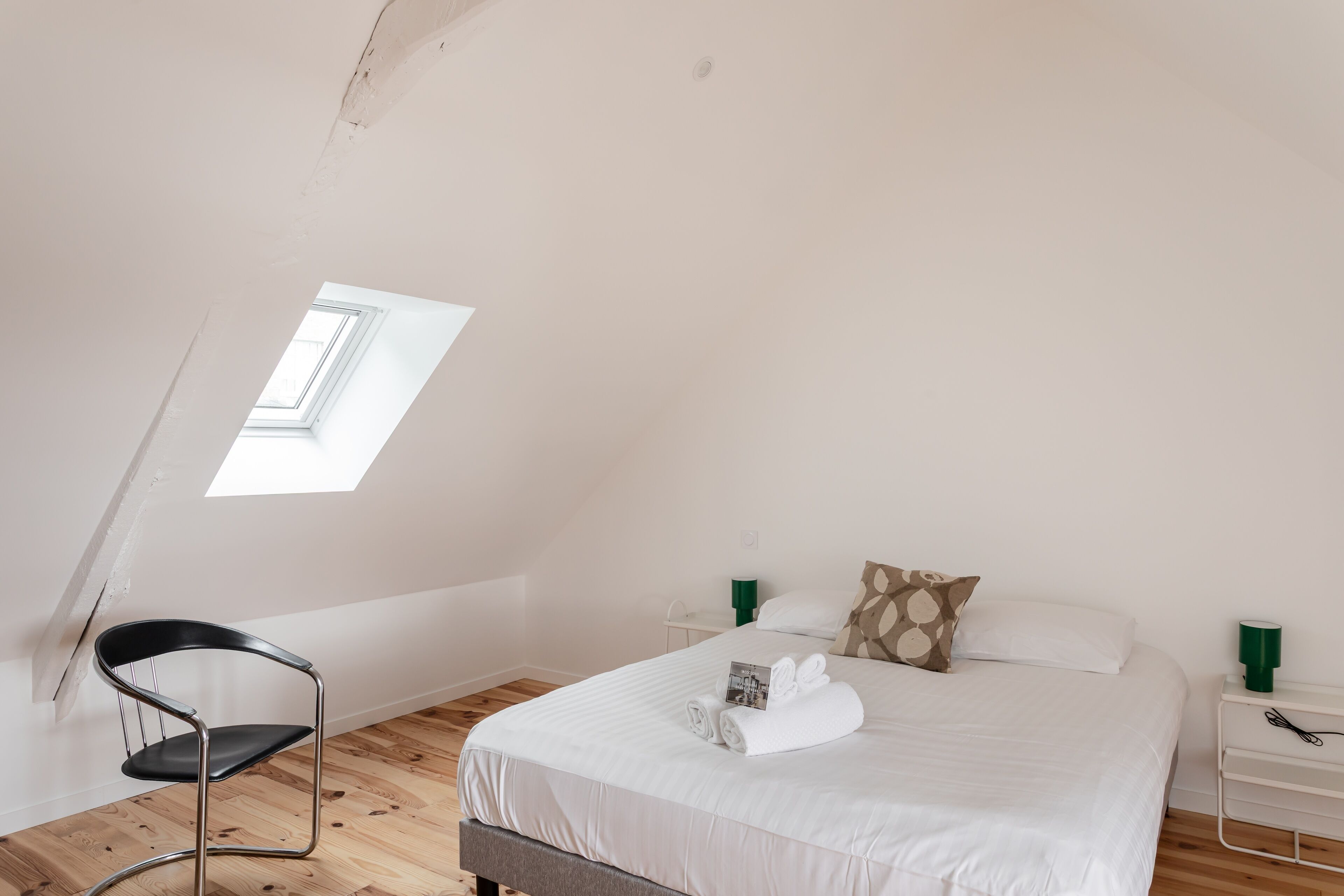 A bedroom with a bed, a chair, a skylight, and a nightstand.