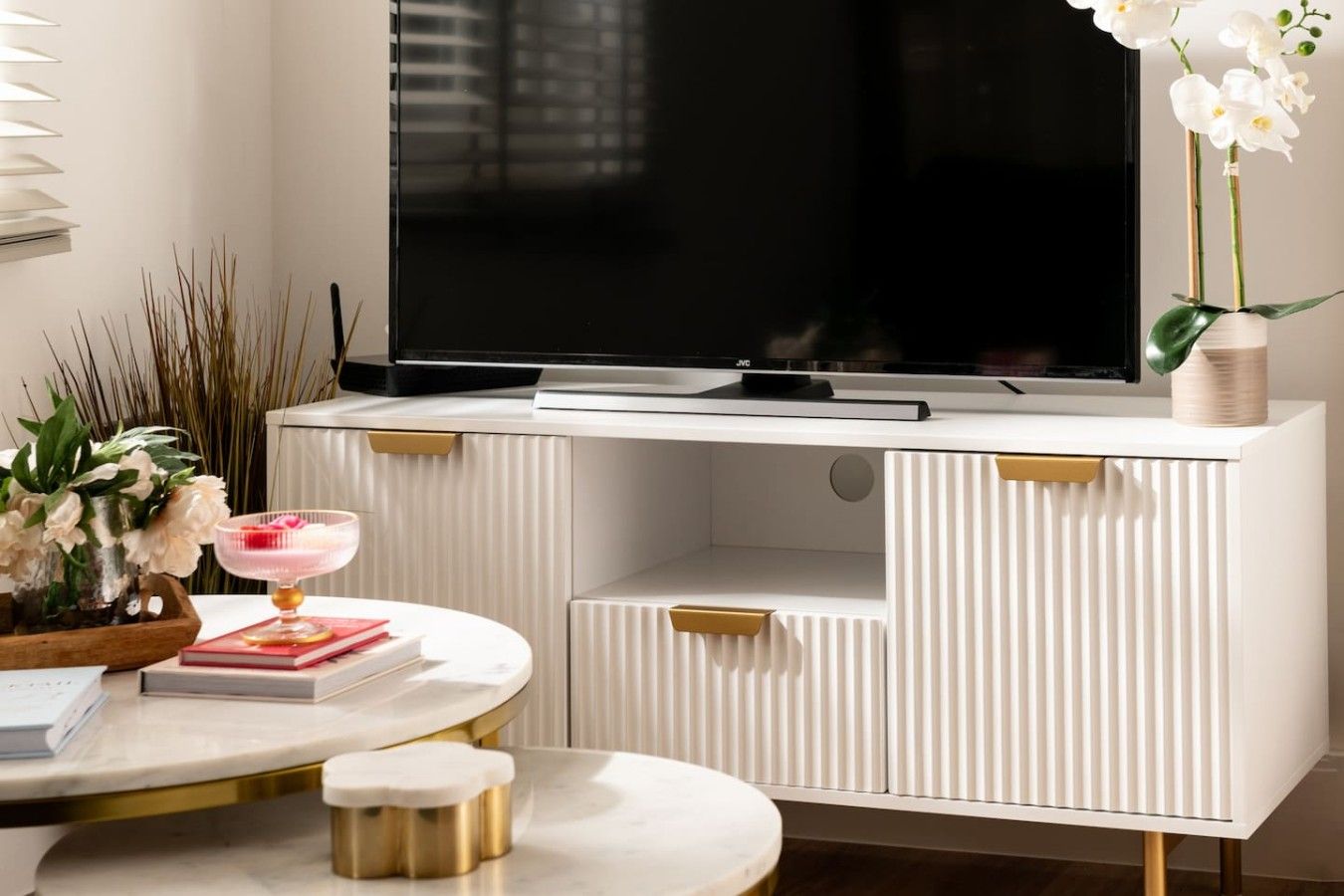 A modern living room with a flat-screen TV, a white entertainment unit, a round coffee table, and decorative plants.