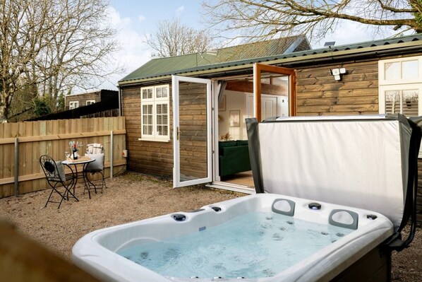 A hot tub in a backyard with a wooden house, a table with chairs, and a covered patio area.