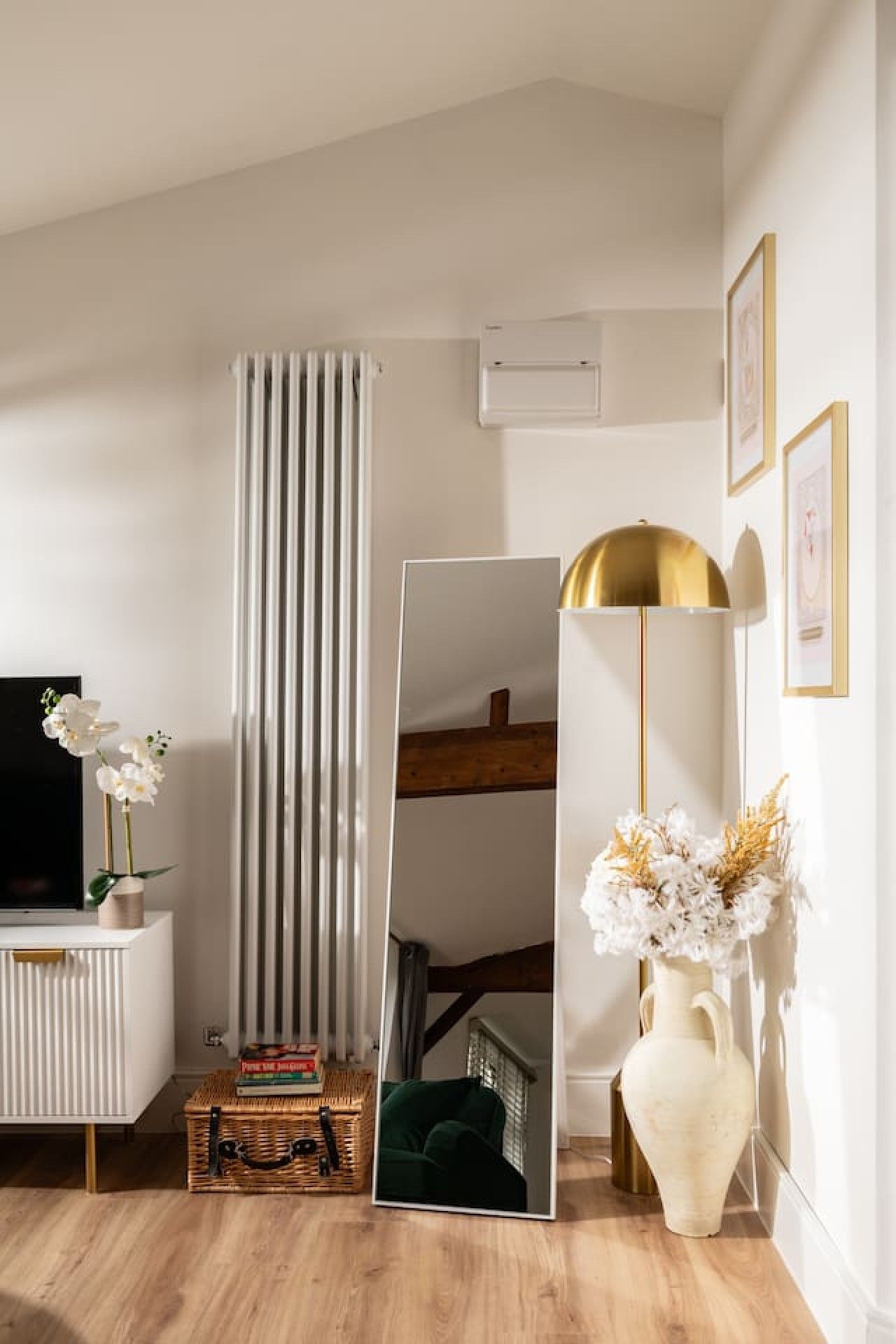 A room with a white radiator, a large mirror, a television, a wooden cabinet, and a vase with flowers.