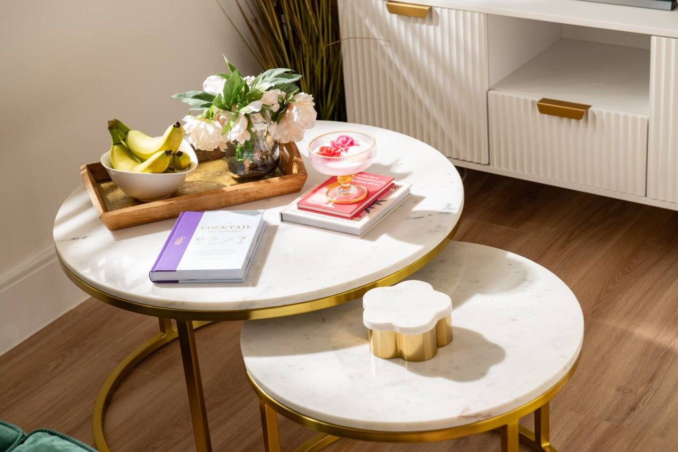 Una mesa redonda con un libro, una bandeja con plátanos, un florero con flores y un pequeño recipiente blanco.