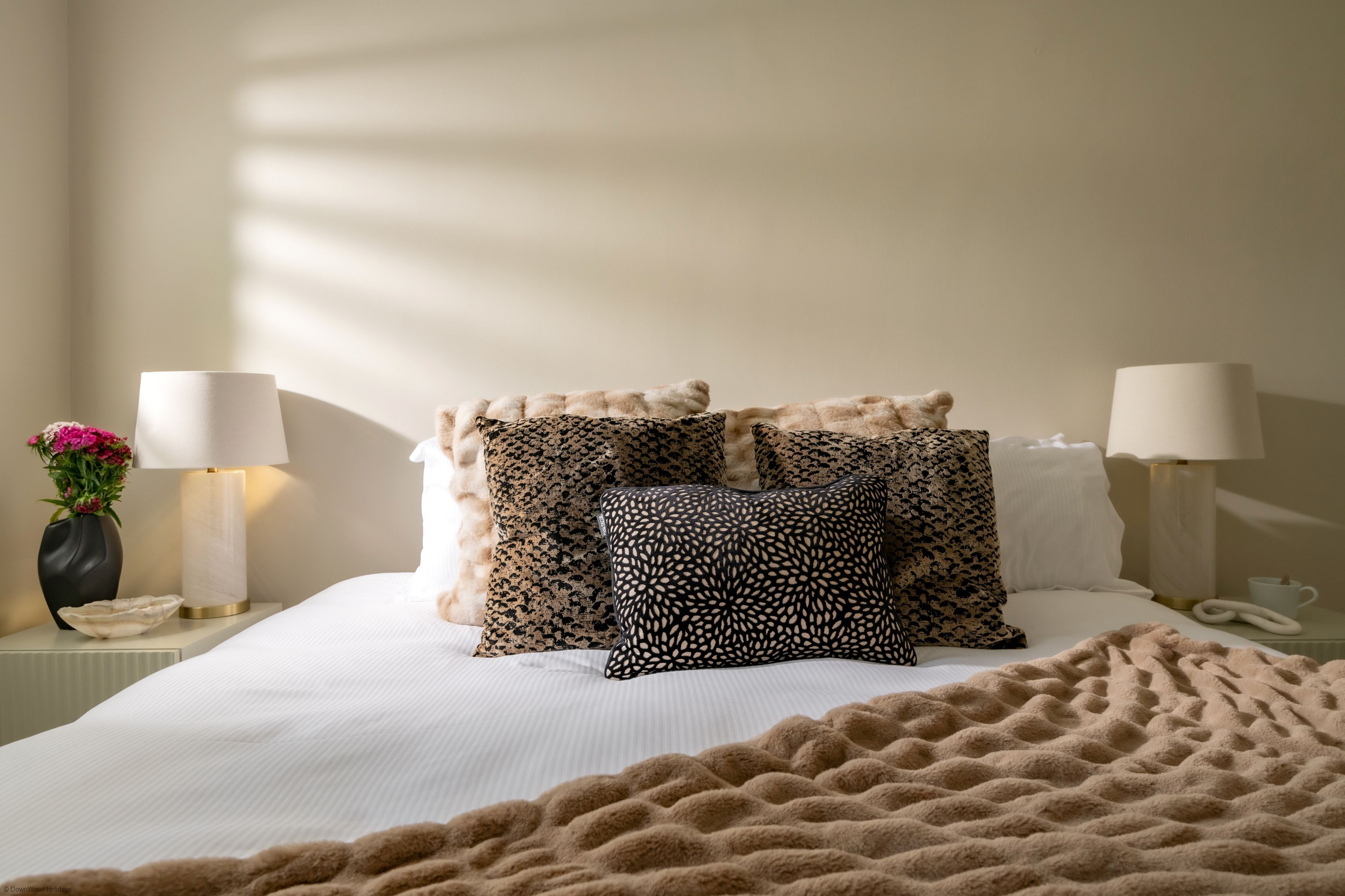 Una cama bien hecha con almohadas estampadas y un edredón esponjoso, un florero con flores y lámparas de noche en una mesita.