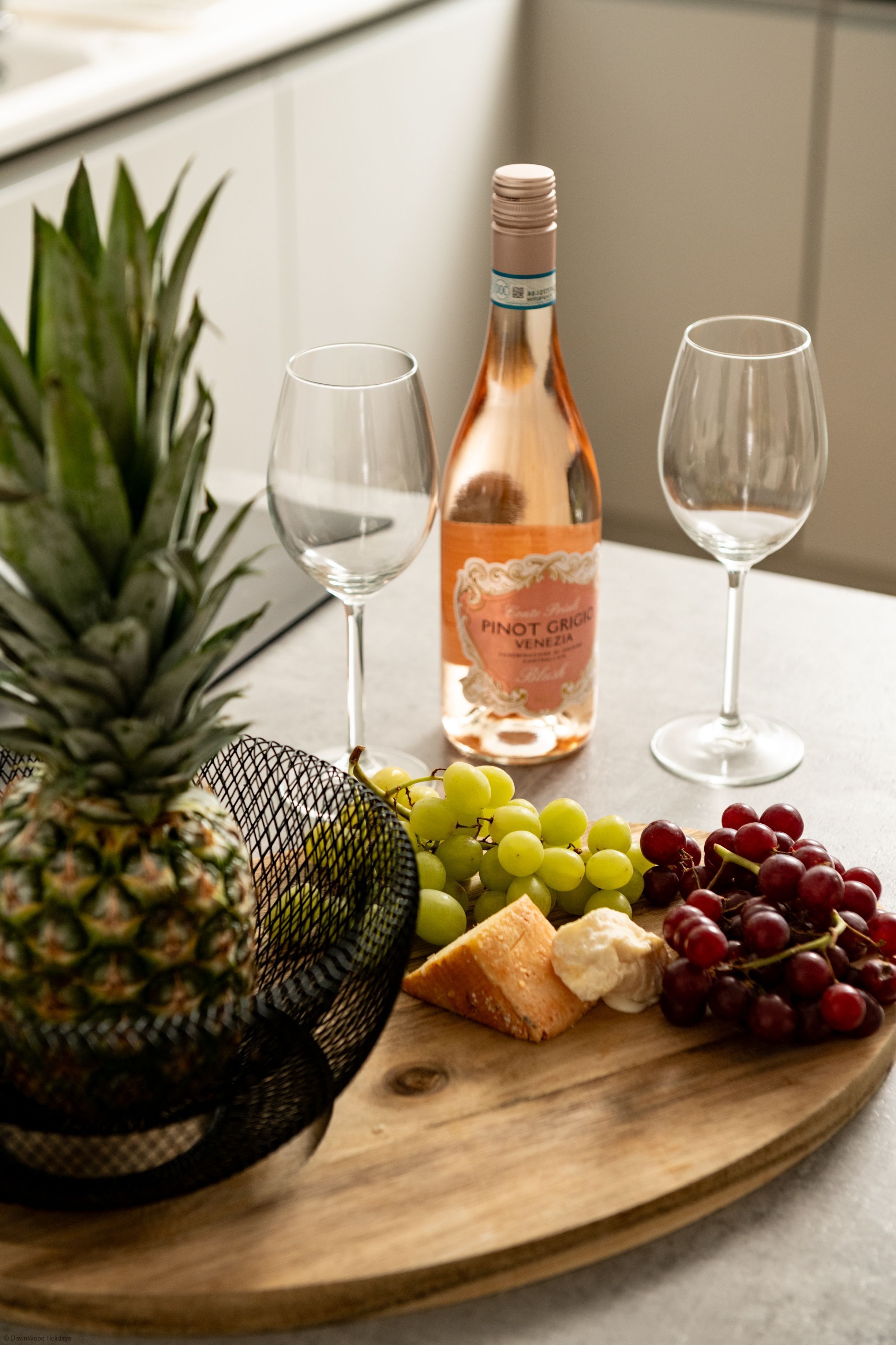 Puinen tarjoilulauta, jonka päällä on pullo Pinot Grigio -viiniä, kaksi viinilasia, ananas, rypäleitä ja juustoa.