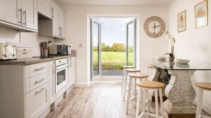Une cuisine avec des meubles blancs, un four à micro-ondes et un appareil à faire du pain. Une porte vitrée donne accès à un patio aménagé avec des meubles d’extérieur et offrant une vue sur la verdure.