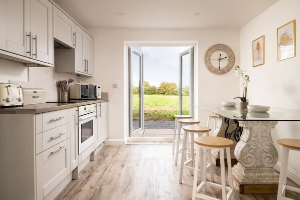 A kitchen with white cabinets, a microwave, and a bread maker. There is a glass door leading to a patio with outdoor seating and a view of greenery.