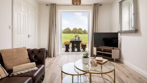 Un salon avec un canapé en cuir, une table basse en verre, un téléviseur à écran plat et une vue sur un champ verdoyant, visible à travers une porte vitrée.