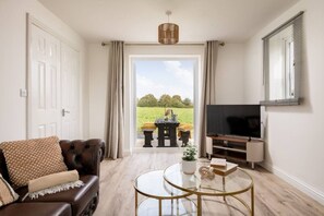 A living room with a leather sofa, a glass coffee table, a flat-screen TV, and a view of a green field through a glass door.