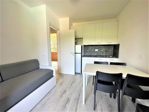 A compact living space with a sofa, a dining table with chairs, a kitchen area with cabinets, and a refrigerator. - Bibione Flat With Pool Access (San Michele al Tagliamento)