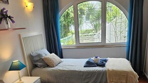 Habitación de hotel con dos camas, una ventana con cortinas, una lámpara de noche y un aplique de pared.