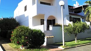 Un edificio blanco de dos pisos con un balcón, un pequeño jardín con flores rojas y una farola.