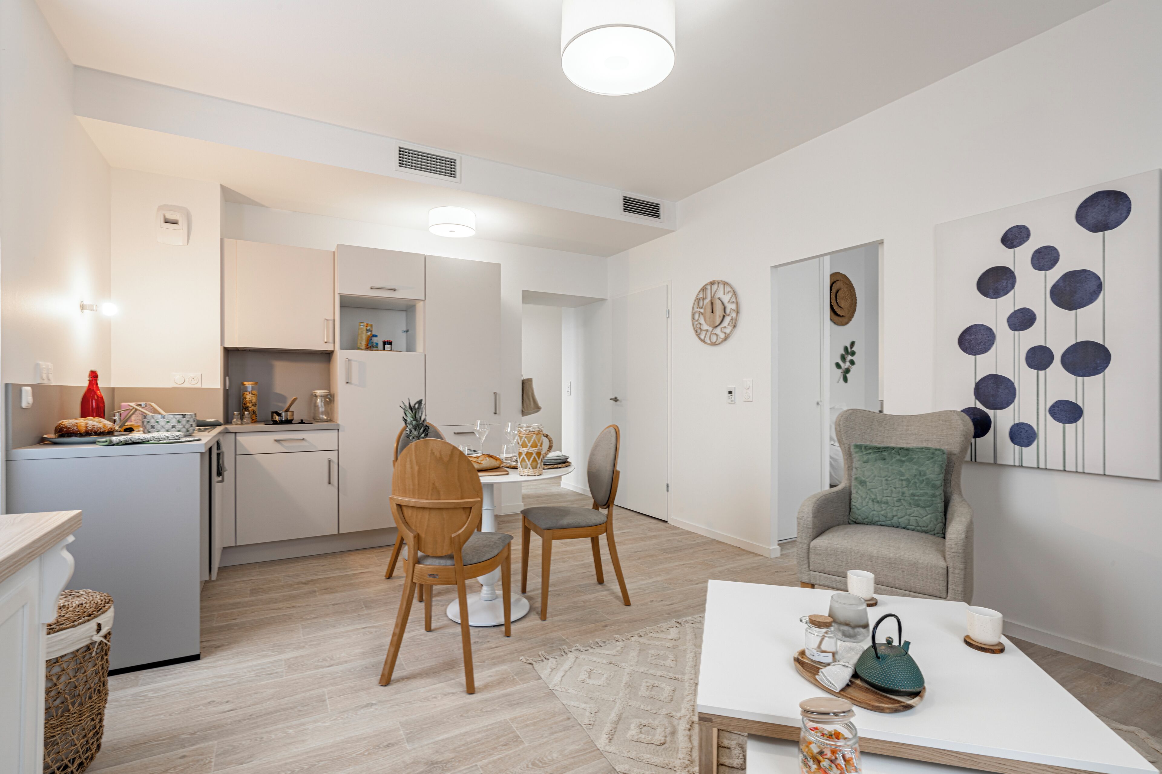 A modern kitchen with white cabinets, a dining area with a white table and two wooden chairs, and a living area with a sofa and a decorative wall piece.