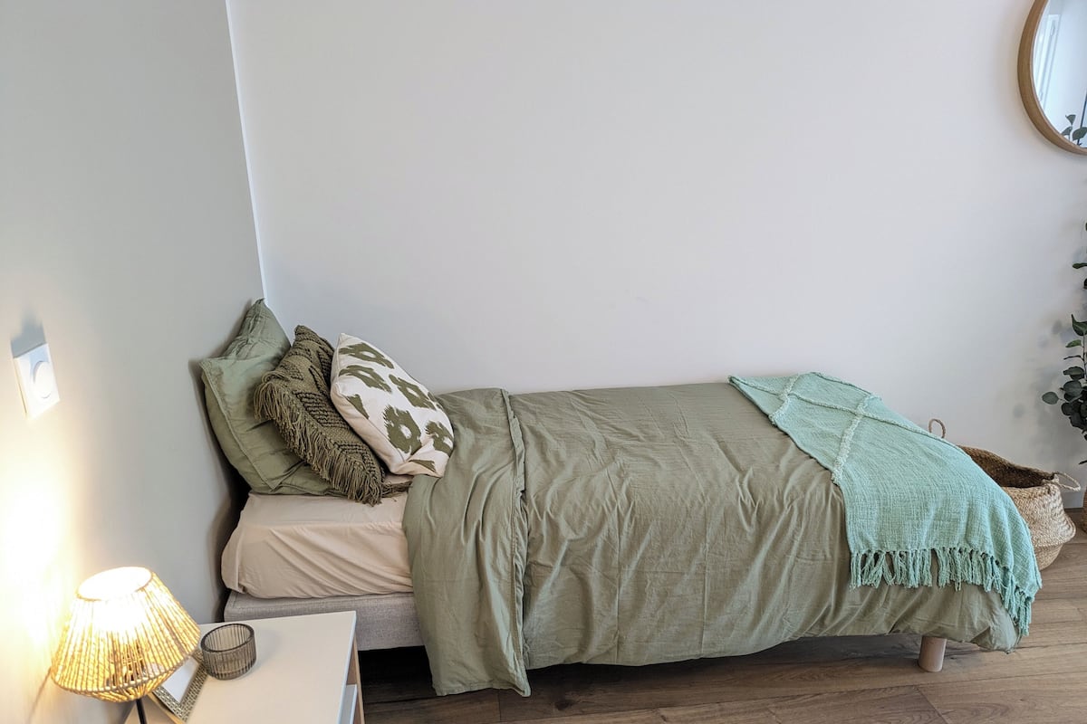 A single bed with a green blanket and two pillows, a bedside table with a lamp, and a wall-mounted mirror.