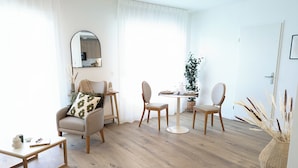 Un salon moderne avec une table à manger ronde, deux chaises, un canapé agrémenté d’un coussin à motifs, une petite table d’appoint et un grand miroir mural.