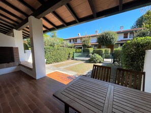 A covered patio area with a wooden table and chairs, a brick fireplace, and a view of a residential neighborhood with houses and greenery. - Great Two-storey Home (San Michele al Tagliamento)