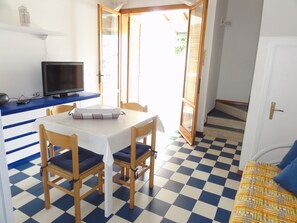 Una habitación con piso a cuadros, una mesa con sillas, un televisor sobre un mueble y una puerta que da al exterior.