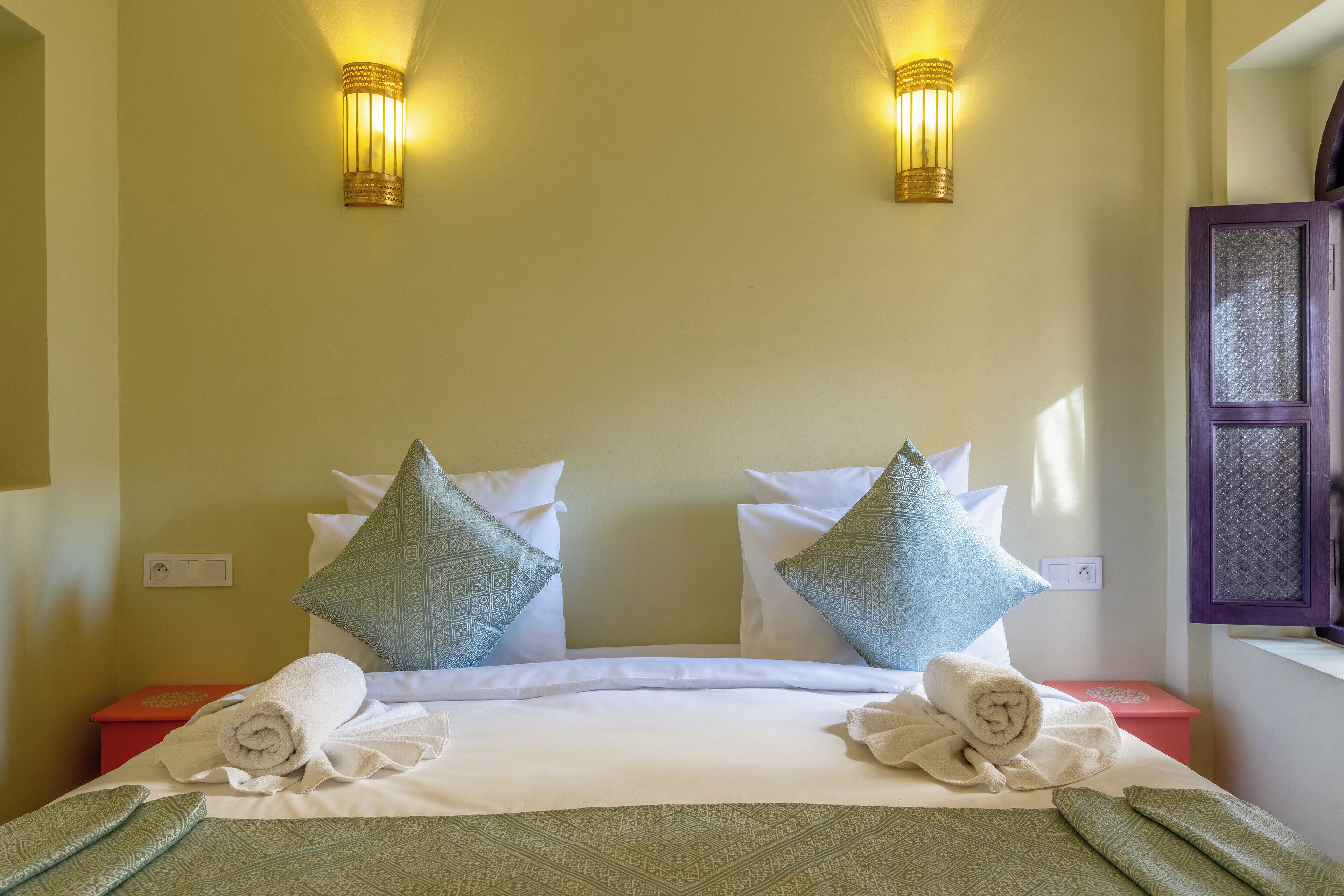 Una cama bien hecha con sábanas blancas, dos almohadas decorativas y toallas enrolladas en el cabecero.