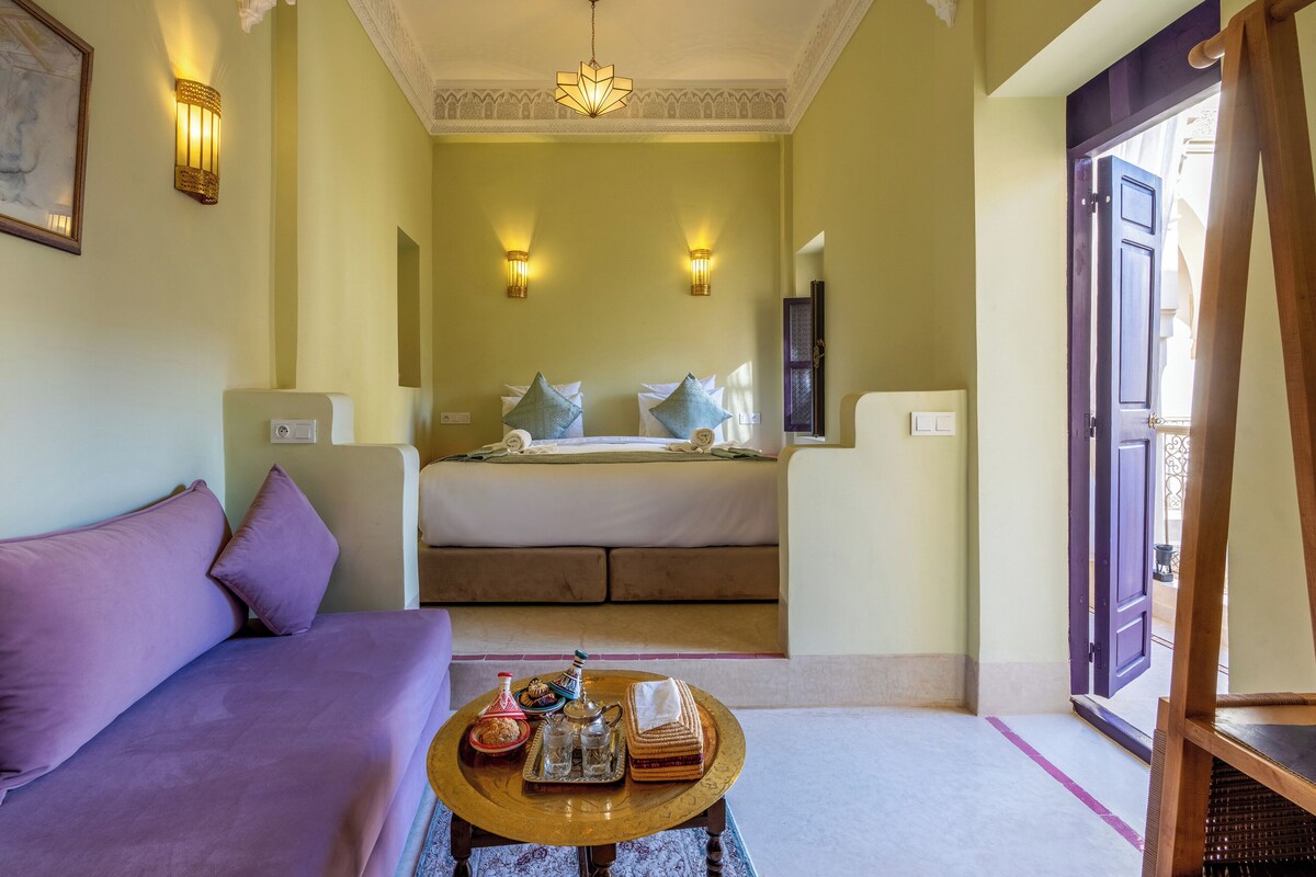A hotel room with a bed, a sofa, a round table with a tray of items, and two wall-mounted lights.