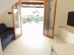 Una habitación con puerta corrediza de vidrio que da a un espacio exterior, un sofá azul oscuro, una mesa blanca y un televisor montado.