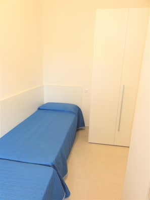 Ein kleines Zimmer mit einem Einzelbett, einem schlichten weißen Wandschrank und einem hellen Boden.