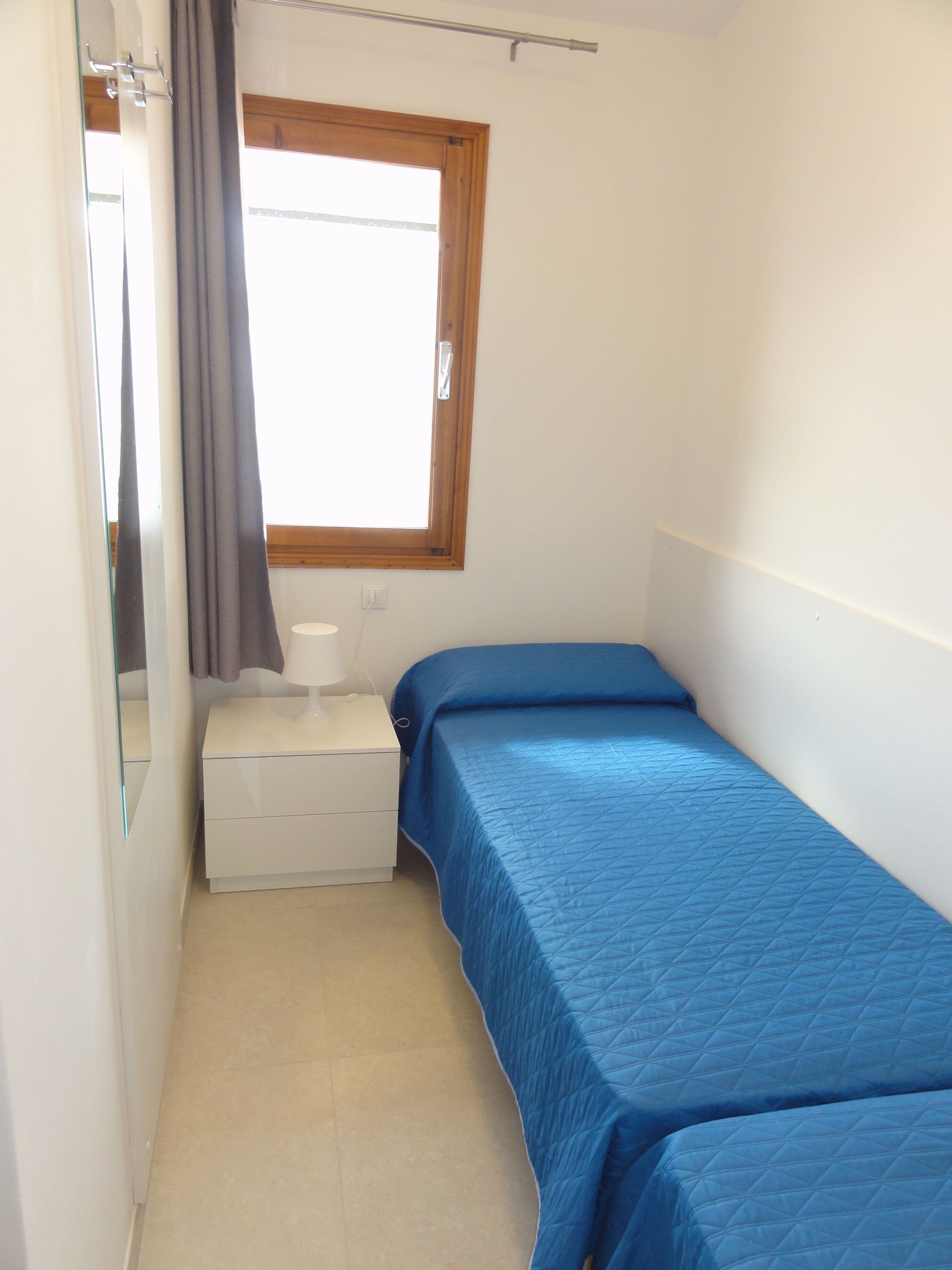 A small, single-bed room with a window, a bedside table, and a mirror.
